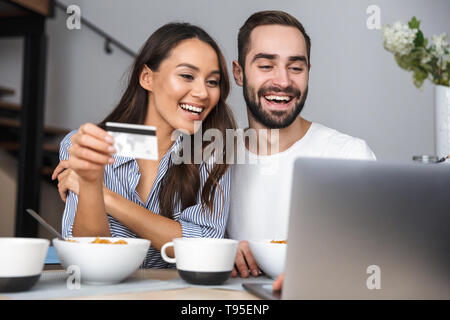 Happy multiethnischen Paar Frühstück in der Küche, am Laptop suchen, mit Kreditkarte Stockfoto