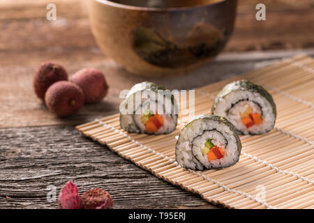 Drei Maki Sushi Rollen auf Bambus Matte auf Holz- Oberfläche, traditionellen asiatischen Tee-Schale, Litschi und Litschis schälen - Schrägansicht, unscharfen Hintergrund, landscap Stockfoto