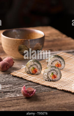 Drei Maki Sushi Rollen auf Bambus Matte auf Holztisch, traditionellen asiatischen Tee-Schale, Litschi und Litschis schälen - gewinkelt, unscharfen Hintergrund, vertikale f Stockfoto