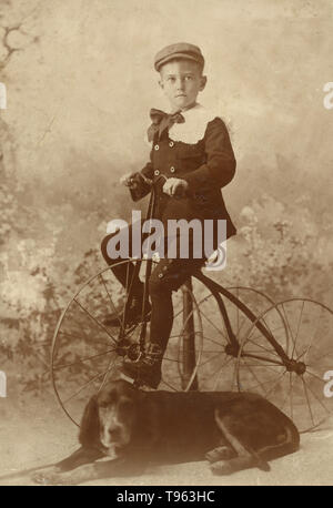 Junge trägt eine Kappe, montiert auf einem alten Stil dreirädrigen Fahrrad, mit einem Hund im Vordergrund, 1890. Von Henry Davis, Amerikanische genommen. Stockfoto