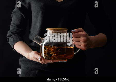 7/8-Ansicht der jungen Frau mit transparenten Teekanne mit chinesische Teeblumen isoliert auf Schwarz Stockfoto