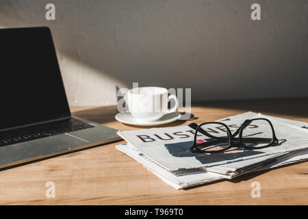 Selektiver Fokus der Brille auf der Wirtschaftszeitung in der Nähe von Laptop und Tasse mit Untertasse auf hölzernen Tisch Stockfoto