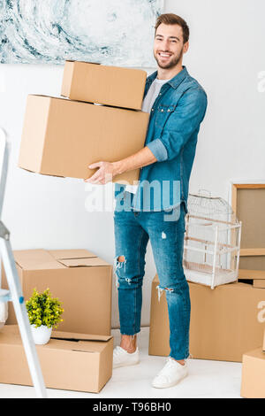 Die volle Länge der lachende Mann in Jeans holding Kartons und Kamera Stockfoto