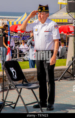 Eine militärische Veteran Mitglied der amerikanischen Legion begrüßt nach dem POW/MIA-Flag auf einen symbolischen leeren Stuhl mit einem Sinnbildlichen T Shirt zu einem Veterans Day Einhaltung in Huntington Beach, CA. Stockfoto