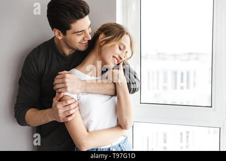 Portrait von positiven Paar lächelnd und zusammen Umarmen beim Stehen in der Nähe von großes Fenster im hellen Zimmer zu Hause Stockfoto