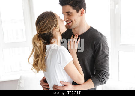 Portrait von europäischen Paar lächelnd und zusammen Umarmen beim Stehen in der Nähe von großes Fenster im hellen Zimmer zu Hause Stockfoto