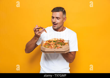 Bild Der unrasierte Mann 30 s im weißen T-Shirt halten und essen Pizza im Stehen über Gelb Hintergrund isoliert Stockfoto