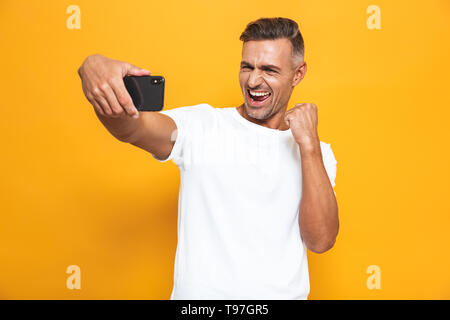 Bild Der unrasierte Mann 30 s im weißen T-Shirt lächelnd und unter selfie Foto auf Mobiltelefon über gelben Hintergrund isoliert Stockfoto