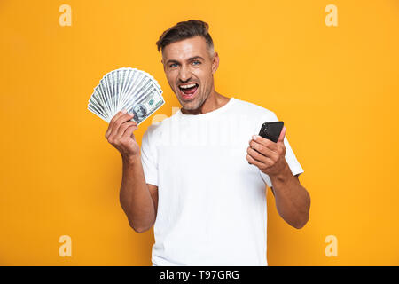 Bild der Unrasierte Kerl 30 s im weißen T-Shirt, Handy und Bündel Geld über Gelb Hintergrund isoliert Stockfoto
