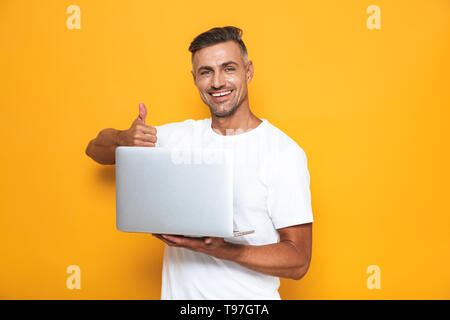 Bild der Unrasierte Kerl 30 s im weißen T-Shirt und mit Silber Laptop über Gelb Hintergrund isoliert Stockfoto