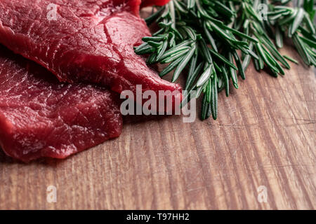 Zwei Stücke von rohem Rindfleisch mit frischem Grün Rosmarin Zweige liegen auf einem Holzbrett mit leeren Raum für Text Stockfoto