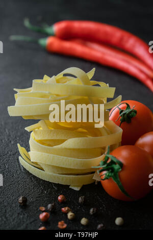 Auf schwarzem Hintergrund Kugeln von Roh Fettuccine Pasta mit frischen Tomaten mit Wurzeln, schwarze und rote Pfefferkörner und zwei Kaffeepads von Chili Stockfoto