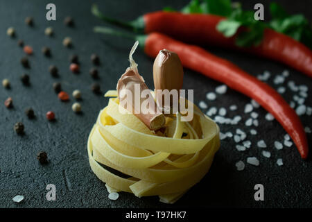 Auf einem dunklen Hintergrund roh Fettuccine einfügen Top zwei Knoblauchzehen, hinter ist Chili, mit grünen Zweigen von Cilantro, grobes Meersalz Schwarz und Stockfoto