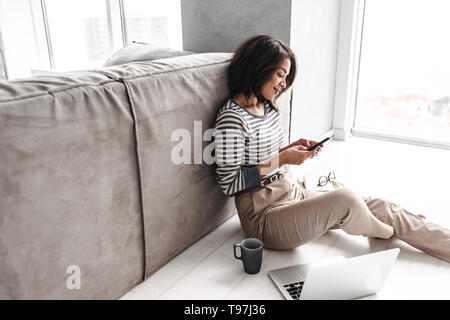 Attraktive junge afro-amerikanische Frau an einem Tisch zu Hause sitzen, Laptop, Kaffee trinken, mit Handy Stockfoto