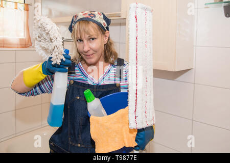 Eine blonde Frau motiviert ist das Bad zu reinigen. Sie trägt ein Kopftuch und Latzhose. In den Händen hält sie Utensilien für die Reinigung. Stockfoto