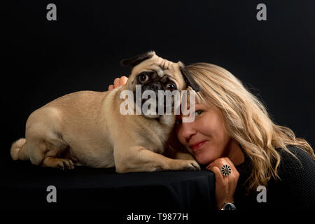 Eine Frau mit langen blonden Haaren Knuddel mit ihrem Hund (PUG). Im Studio, das blonde Haar glänzt wunderschön vor schwarzem Hintergrund. Stockfoto