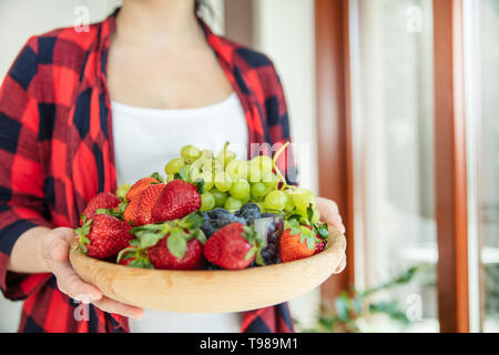 Frau hält hölzerne Schüssel mit grünen Trauben, Erdbeeren und Blaubeeren mit Fenster im Hintergrund. Stockfoto
