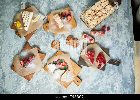 Verschiedene süße Desserts: Kuchen, churos, Baiser roll, Eis, mit Beeren auf Holz dekoriert. Ansicht von oben. Grau strukturierten Hintergrund. Schöne Gerichte. Nachtisch. Lebensmittelkette Stockfoto
