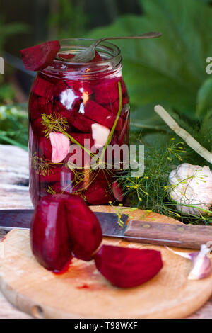 Hausgemachte Marmeladen, Eingelegte Rüben in einem Glas auf einem Holztisch unter Kräuter und Gemüse, Knoblauch. Rote-bete-Schnitt auf ein Holzbrett. Stockfoto