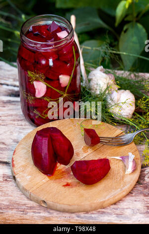 Hausgemachte Marmeladen, Eingelegte Rüben in einem Glas auf einem Holztisch unter Kräuter und Gemüse, Knoblauch. Rote-bete-Schnitt auf ein Holzbrett. Stockfoto