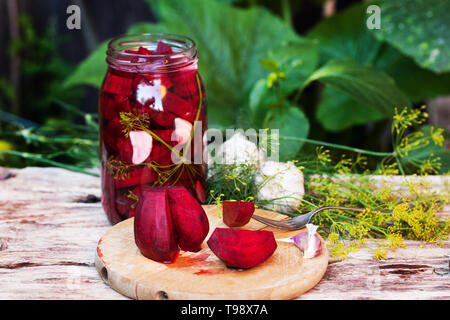 Hausgemachte Marmeladen, Eingelegte Rüben in einem Glas auf einem Holztisch unter Kräuter und Gemüse, Knoblauch. Rote-bete-Schnitt auf ein Holzbrett. Stockfoto