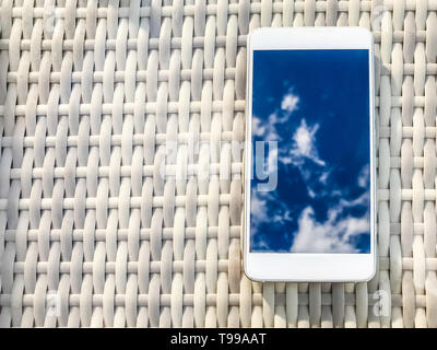 Reflexion der blauen Himmel mit Wolken im Telefon - Smartphone, das auf einem hellen Rattanmöbeln Tabelle vor dem Hintergrund liegt. Blur und Abstraktion Stockfoto
