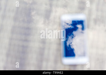 Reflexion der blauen Himmel mit Wolken im Telefon - Smartphone, das auf einem hellen Rattanmöbeln Tabelle vor dem Hintergrund liegt. Blur und Abstraktion Stockfoto