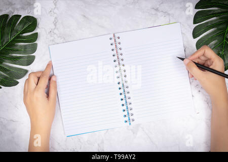 Flach der Frau Hand schreiben in eine leere weiße Seite notebook auf weißem Marmor Schreibtisch, Ansicht von oben. Green Palm Leaf auf dem Schreibtisch für die Dekoration Stockfoto