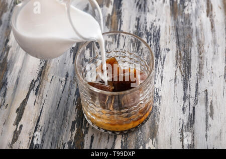 Das Gießen von Milch in Glas mit Kaffee Eiswürfel auf hölzernen Tisch Stockfoto