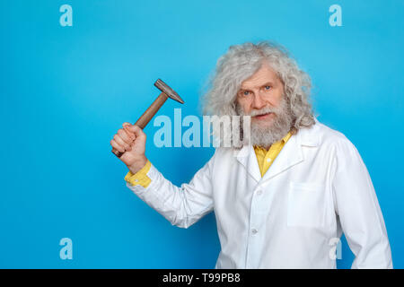 Alte langhaarigen Mann, der Doctor's Kleid Studio auf der blauen Wand holding Hammer suchen Kamera isoliert Ernst Stockfoto