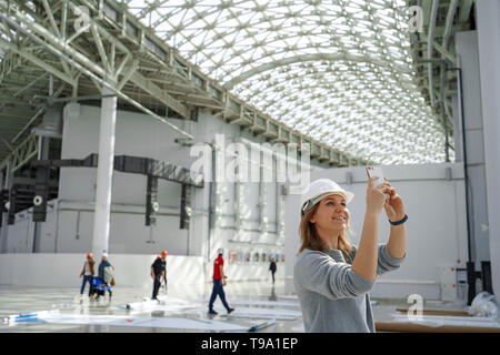 Junge Mädchen in einem Helm nimmt Bilder auf dem Handy, im Hintergrund der Arbeitnehmer, eine Exkursion zur Baustelle, ein Ingenieur ist zufrieden Stockfoto