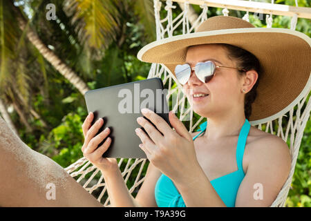 Lächelnde junge Frau mit Sonnenbrille und Hut mit digitalen Tablet liegen auf Hängematte am Strand Stockfoto