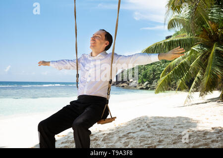 Lächelnden jungen Mann sitzt auf Swing Outstretching seine Hand geniessen die frische Luft am Strand Stockfoto