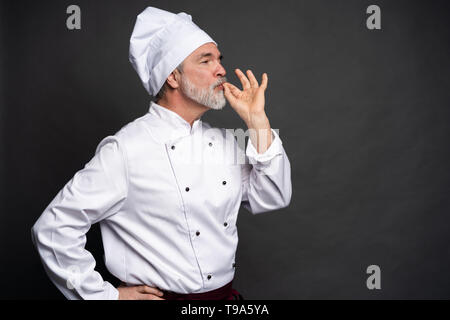 Koch, lecker lecker Geste durch Küssen Finger. Zuversichtlich bärtigen Mann Koch in weiße Uniform mit perfekter unterzeichnen. Stockfoto