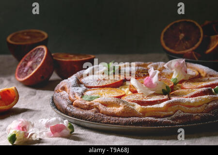 Hausgemachter Käsekuchen mit sizilianischer Blutorangen, dekoriert von essbaren Blüten, Minze und Zucker Pulver in der Platte mit geschnittenen Orangen oben g serviert. Stockfoto