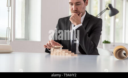 Ernste nachdenkliche Geschäftsmann in seinem Büro Schreibtisch Montage eine Treppe aus Holz sitzen Pflöcke in einem konzeptionellen Bild des Unternehmens Vision und Ehrgeiz. Stockfoto