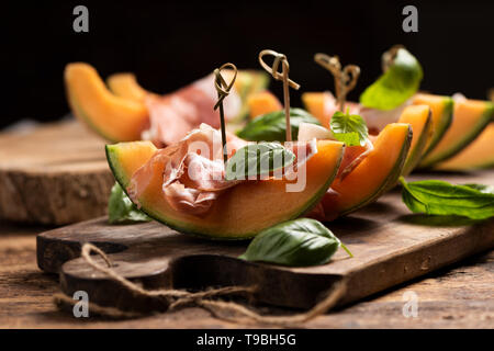 Aufgeschnittene Melone mit Schinken und Basilikum, serviert auf einem Holz Schneidebrett Nahaufnahme Stockfoto