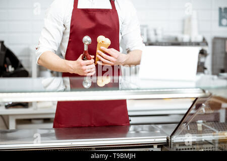 Verkäufer halten Eis in der Waffel Kegel und professionelle Schaufel auf die rote Schürze, Hintergrund, Nahaufnahme Stockfoto