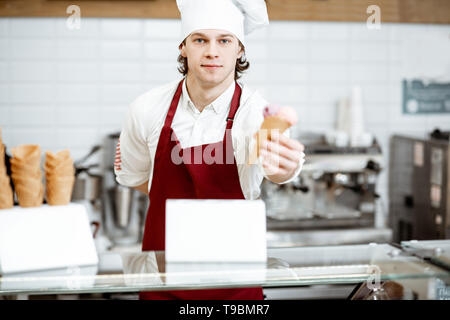 Junge Küchenchef Koch oder Verkäufer in Schürze und hat den Verkauf von Eis in der Waffel Kegel in der modernen Konditorei Stockfoto