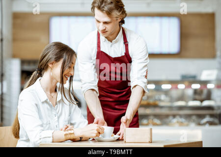 Stattliche Kellner in rot Schürze mit süßer Kuchen für eine junge Frau, die Kunden bei der Konditorei und Café Stockfoto