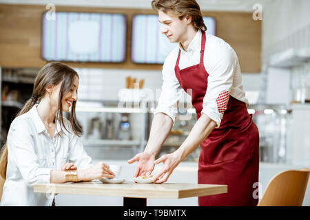 Stattliche Kellner in rot Schürze mit süßer Kuchen für eine junge Frau, die Kunden bei der Konditorei und Café Stockfoto