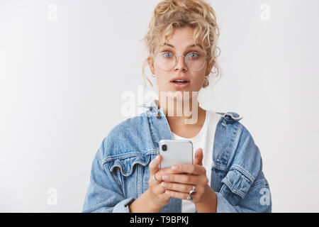 Fragte mich erstaunt cute blonde Frau unordentlich Curly bun Brille trägt Jeansjacke spielen coolen Smartphone Spiel holding Telefon Adresse suchen mit Telefon Stockfoto