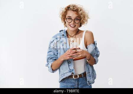 Stilvolle und glücklich nette charismatische weibliche Mode blogger in Gläsern mit kurzen blonden Haare schneiden Lächeln breit Holding Smartphone unter Foto Stockfoto