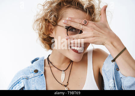 Peekaboo, die dort. Porträt des charismatischen niedlich und verspielt Freundin mit stilvollen curly Frisur, Brille und Jeansjacke schließen Augen mit Stockfoto