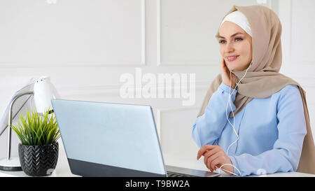 Porträt der jungen muslimischen Frau im hijab und traditionelle Musik hören ist der Verschleiß der Ohrhörer mit Laptop und es geniessen Sitzen am Tisch. Stockfoto