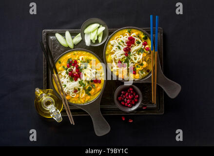 Laksa mit Kürbis und Kokosmilch, Reisnudeln, Brokkoli und Granatapfel Samen portioniert in die schwarzen Platten auf einem dunklen Hintergrund. gesund vegan Gericht Stockfoto