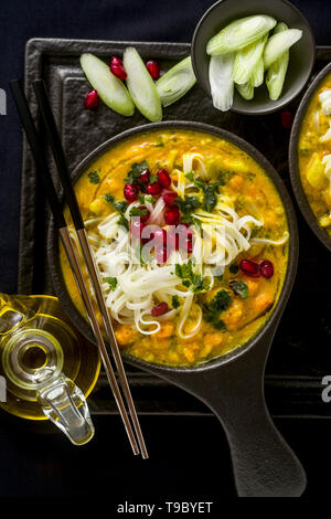 Laksa mit Kürbis und Kokosmilch, Reisnudeln, Brokkoli und Granatapfel Samen portioniert in die schwarzen Platten auf einem dunklen Hintergrund. gesund vegan Gericht Stockfoto