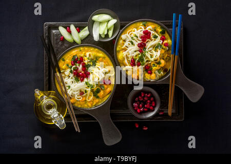 Laksa mit Kürbis und Kokosmilch, Reisnudeln, Brokkoli und Granatapfel Samen portioniert in die schwarzen Platten auf einem dunklen Hintergrund. gesund vegan Gericht Stockfoto