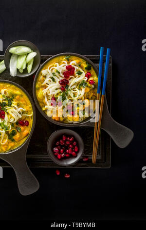 Laksa mit Kürbis und Kokosmilch, Reisnudeln, Brokkoli und Granatapfel Samen portioniert in die schwarzen Platten auf einem dunklen Hintergrund. gesund vegan Gericht Stockfoto