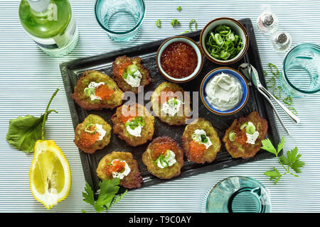 Russische traditionelle Kartoffelpfannkuchen serviert mit rotem Kaviar, grüne Zwiebeln und saurer Sahne auf einem Tablett mit Weißwein und Zitrone Stockfoto